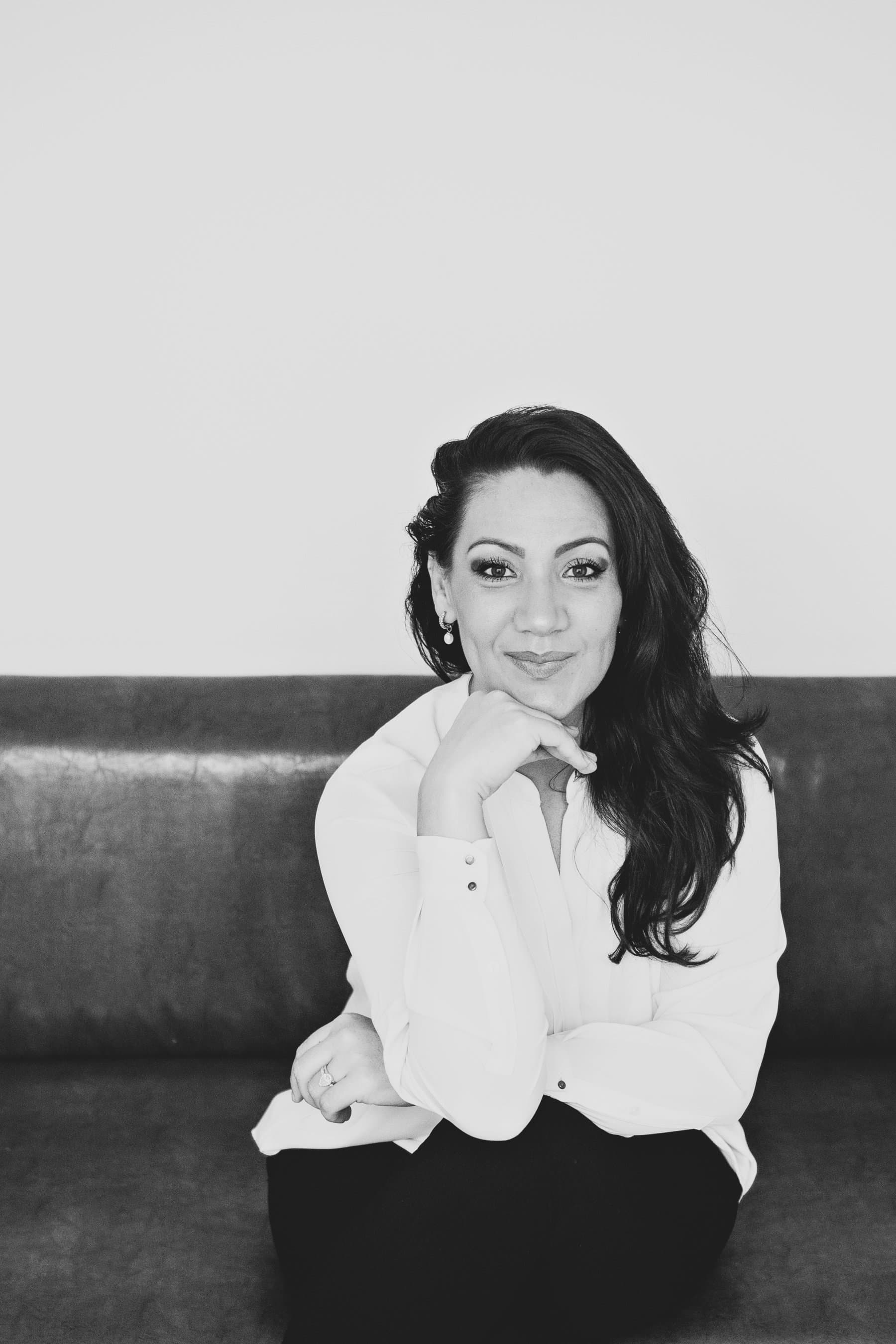 A woman with long dark hair in a white blouse, resting her chin on her hand and smiling